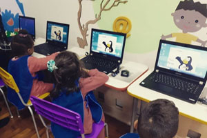 Crianças de frente para computadores com imagens coloridas, e paredes pintadas com imagens de crianças